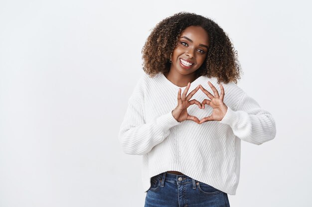 Jolie femme afro-américaine idiote coiffure afro montrer geste coeur tête inclinée souriant joyeusement exprimer la sympathie passion attitude romantique, debout mur blanc insouciant