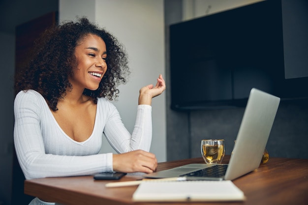Jolie femme afro-américaine souriante assise devant un ordinateur avec du thé et travaillant