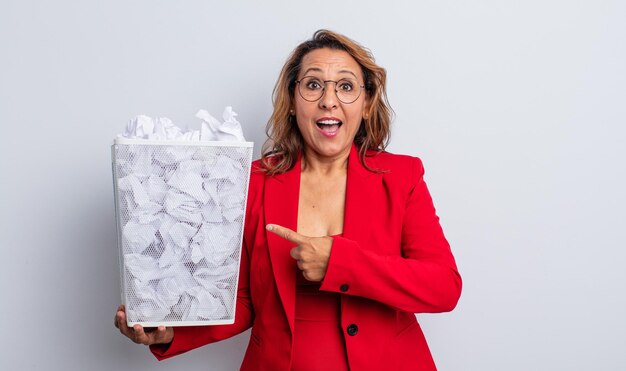 Jolie femme d'âge moyen à l'air excité et surpris en pointant sur le côté. concept de bac à boules de papier