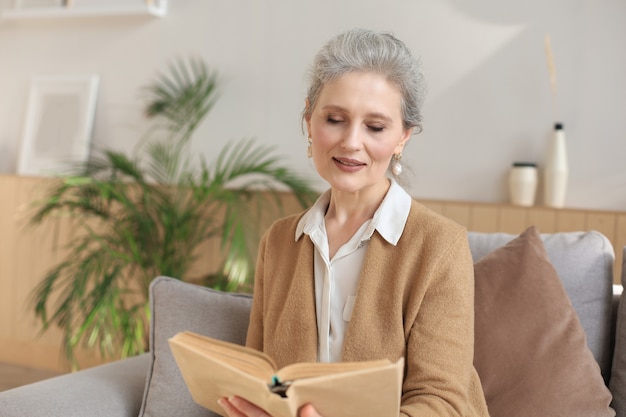 Jolie femme d'âge moyen appréciant la lecture d'un livre assis sur le canapé dans son salon en souriant pendant qu'elle lisait.