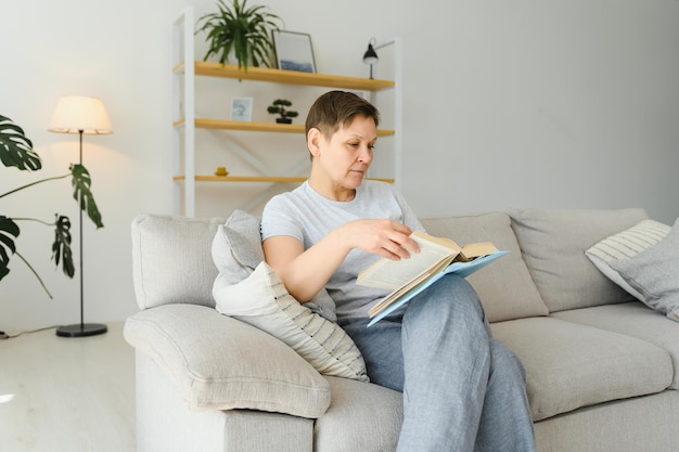 Jolie femme d'âge moyen avec un beau sourire assis sur un canapé dans le salon tenant un livre