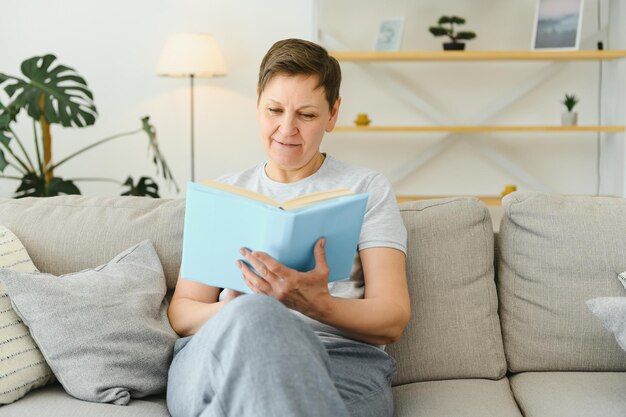 Jolie femme d'âge moyen avec un beau sourire assis sur un canapé dans le salon tenant un livre