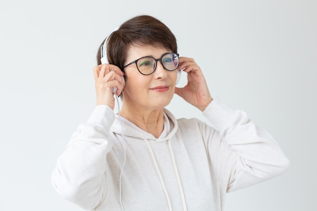 Jolie femme d'âge moyen positive en pull et lunettes écoute de la musique avec des écouteurs en fil debout sur un mur blanc. Concept de loisirs et d'abonnements à la station de radio préférée.