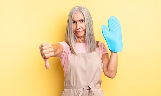 Jolie femme d'âge moyen se sentant croisée, montrant les pouces vers le bas. concept de gants de cuisine