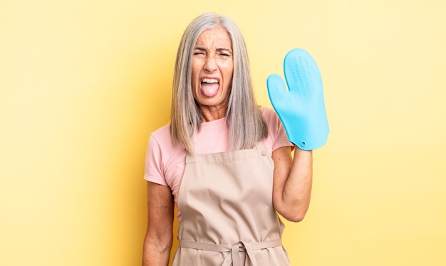 Jolie femme d'âge moyen se sentant dégoûtée et irritée et tirant la langue. concept de gants de cuisine