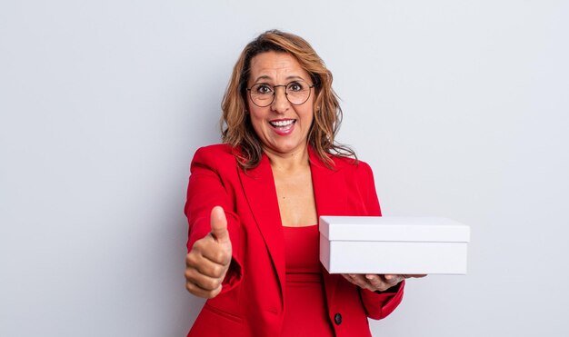 Jolie femme d'âge moyen se sentant fière, souriante positivement avec le pouce levé. paquet de boîte blanche