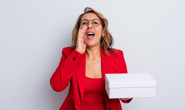 Jolie femme d'âge moyen se sentant heureuse, donnant un grand cri avec les mains à côté de la bouche. paquet de boîte blanche