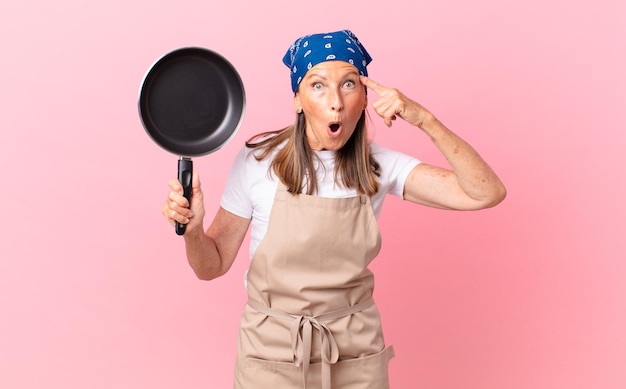 Jolie femme d'âge moyen semblant surprise, réalisant une nouvelle pensée, idée ou concept et tenant une casserole. concept de chef