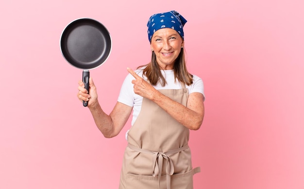 Jolie femme d'âge moyen souriant joyeusement, se sentant heureuse et pointant sur le côté et tenant une casserole. concept de chef