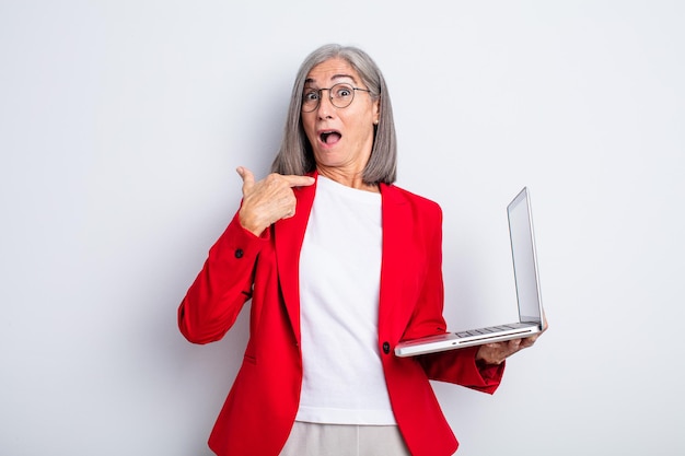 Jolie femme âgée à l'air choquée et surprise avec la bouche grande ouverte, pointant vers soi. concept d'entreprise et d'ordinateur portable