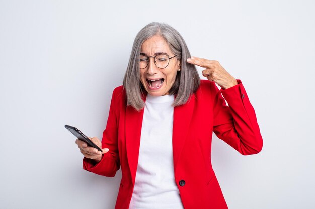 Jolie femme âgée à l'air malheureuse et stressée, geste de suicide faisant signe d'arme à feu. concept d'entreprise et de téléphone