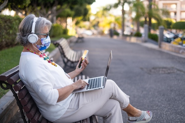 Jolie femme âgée assise sur un banc de parc à l'aide d'un téléphone portable et d'un ordinateur portable profitant de la technologie
