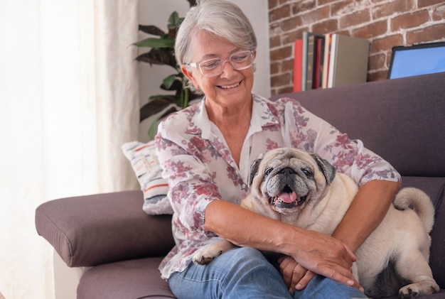 Jolie femme âgée assise sur un canapé à la maison embrassant son vieux chien carlin allongé sur ses jambes