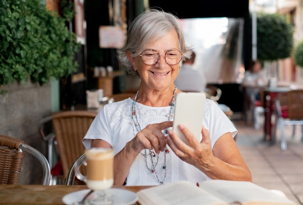 Jolie femme âgée assise à l'extérieur à une table de café en dégustant du café et une boisson au lait tout en utilisant le téléphone caucasienne dame âgée avec des lunettes détendue à la retraite ou en vacances