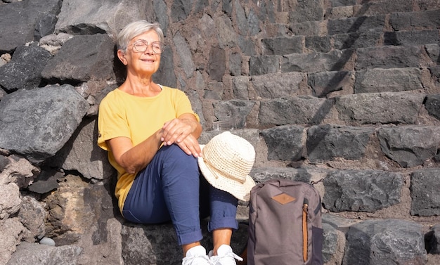 Jolie femme âgée assise les yeux fermés sur un escalier en pierre prenant un bain de soleil Une femme mûre sourit assise près de son sac à dos concept de liberté et de retraite heureuse