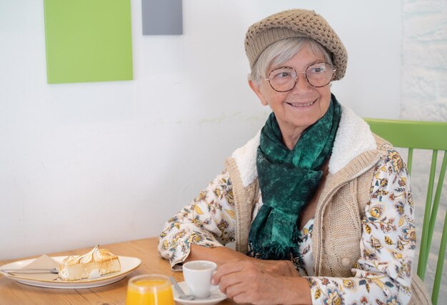 Jolie femme âgée avec chapeau et lunettes assis à la table du café-bar en buvant une tasse de café Petit-déjeuner sur la table avec du jus d'orange et du gâteau