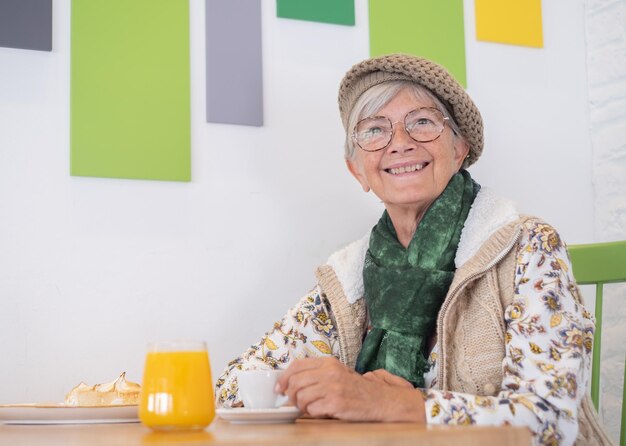 Jolie femme âgée avec chapeau et lunettes assis à la table du café-bar en buvant une tasse de café Petit-déjeuner sur la table avec du jus d'orange et du gâteau