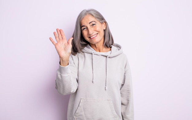 Jolie femme âgée souriante joyeusement et joyeusement, agitant la main, vous accueillant et vous saluant, ou vous disant au revoir