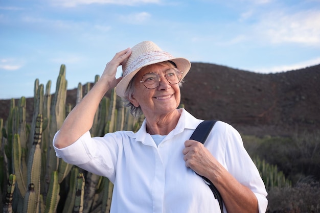 Jolie femme âgée tenant un chapeau en excursion en plein air au coucher du soleil portant un chapeau