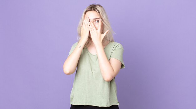 Jolie femme albinos couvrant le visage avec les mains, regardant entre les doigts avec une expression surprise et regardant sur le côté