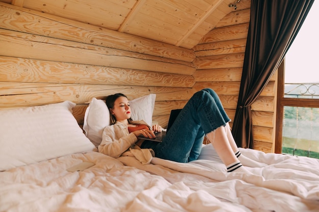 Jolie femme allongée sur le lit dans un appartement avec intérieur en bois et utilisant un ordinateur portable