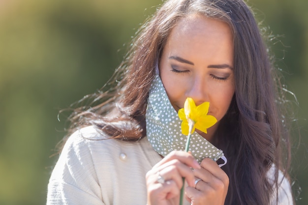 Jolie femme apprécie l'odeur d'une fleur avec la moitié de son masque facial, ayant les yeux fermés par une journée ensoleillée dans la nature.