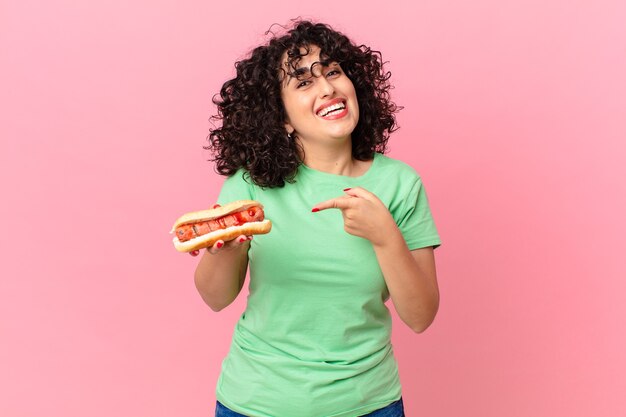 Jolie femme arabe à l'air excité et surpris pointant sur le côté et tenant un hot-dog