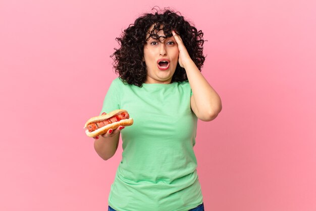 Jolie femme arabe ayant l'air heureuse, étonnée et surprise et tenant un hot-dog