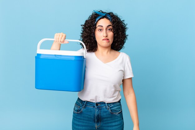 Jolie femme arabe à la perplexité et à la confusion et tenant un réfrigérateur portable