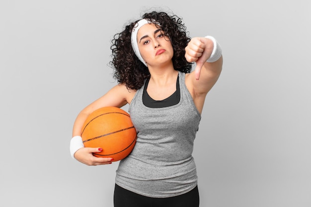 Jolie femme arabe se sentant croisée, montrant les pouces vers le bas et tenant un ballon de basket. notion de sport