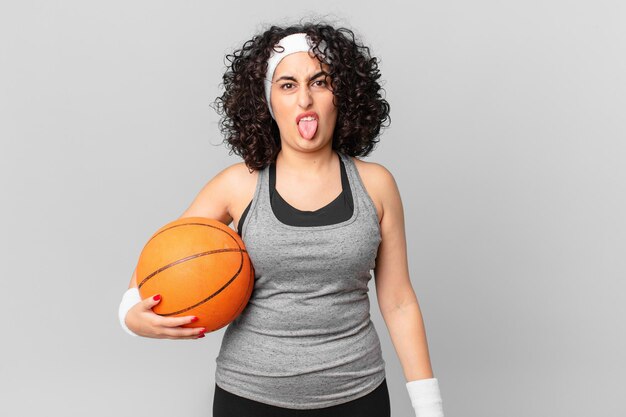 Jolie femme arabe se sentant dégoûtée et irritée et tirant la langue et tenant un ballon de basket. notion de sport
