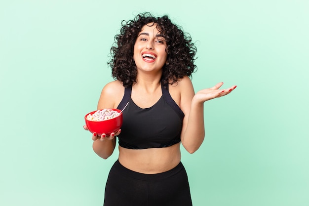 Jolie femme arabe se sentant heureuse, surprise de réaliser une solution ou une idée portant des vêtements de fitness et tenant un bol de flocons de maïs