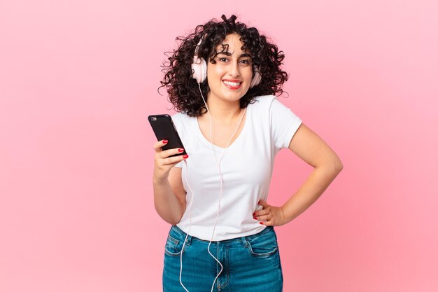 Jolie femme arabe souriant joyeusement avec une main sur la hanche et confiante avec des écouteurs et un smartphone