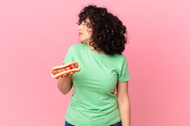 Jolie femme arabe sur la vue de profil pensant, imaginant ou rêvant et tenant un hot-dog