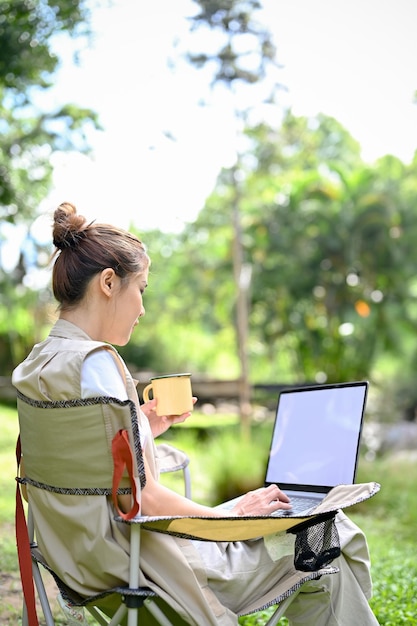 Jolie femme asiatique assise sur la chaise de camping en sirotant un café et en utilisant un ordinateur portable