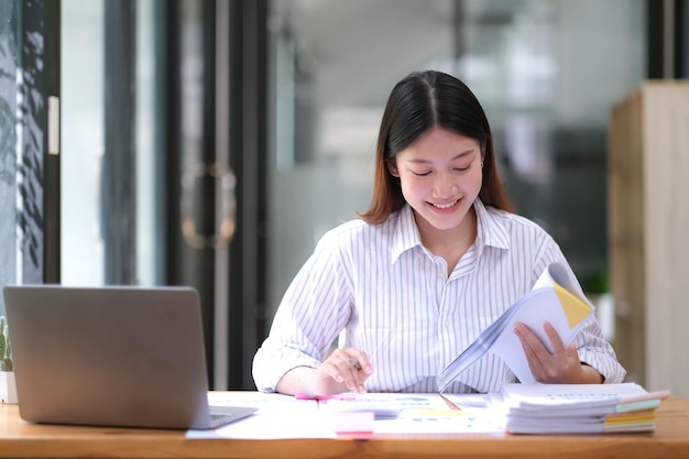 Jolie femme asiatique assise dans la calculatrice de bureau travaillant sur des tableaux de données et des documents sur la table dans l'espace de travail d'entreprise