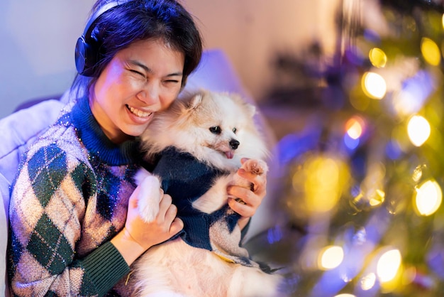 Jolie femme asiatique écouter de la musique de la main du casque câlin mignon petit ami chien de poche avec soin et amour tendresse moment joyeux femme asiatique tenir petit animal de compagnie sur le canapé avec arbre de décoration de noël