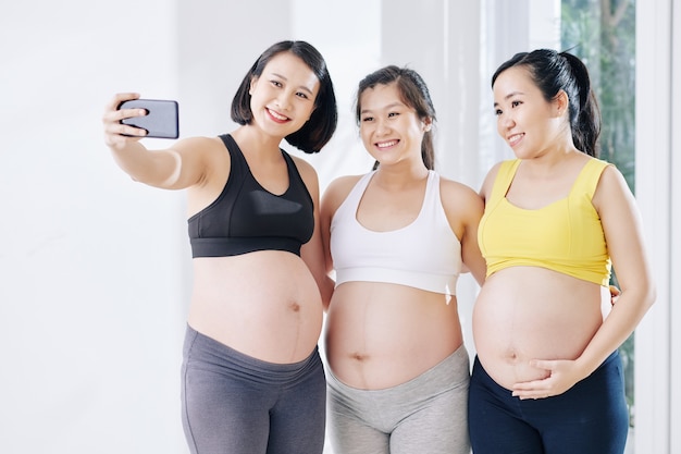 Jolie femme asiatique enceinte souriante prenant un selfie avec d'autres femmes enceintes après un cours de yoga