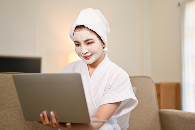 Jolie femme asiatique en peignoir à l'aide d'un ordinateur portable et d'un masque facial à l'argile sur son visage à la maison