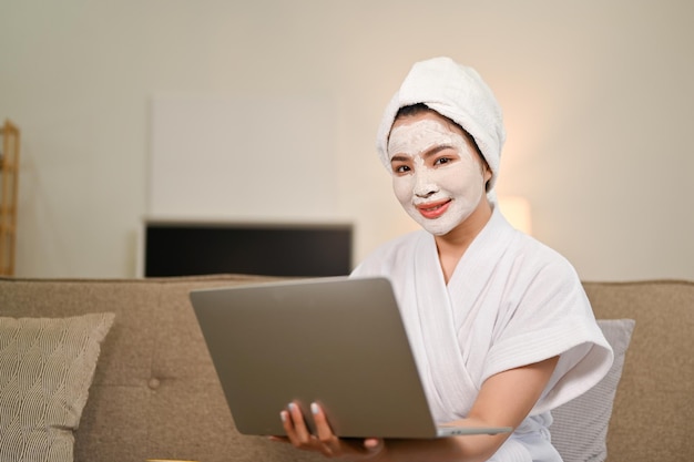 Jolie femme asiatique en peignoir à l'aide d'un ordinateur portable tout en appliquant un masque de traitement facial à la maison