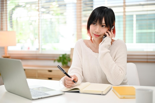 Jolie femme asiatique prenant des notes tout en parlant au téléphone avec son client