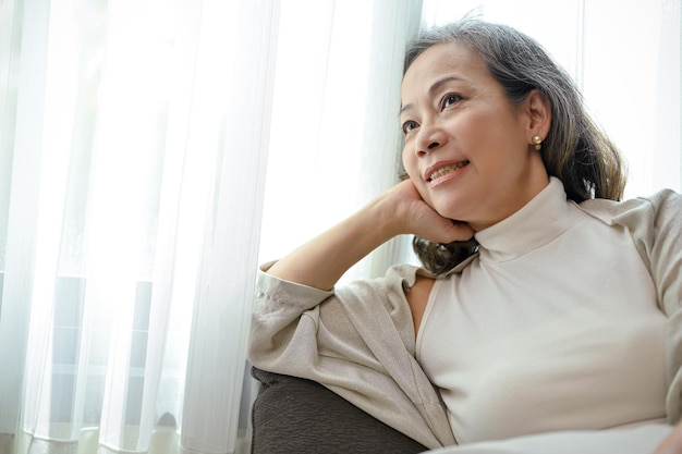 Jolie femme asiatique se détend dans le salon est assis sur un canapé en regardant par la fenêtre