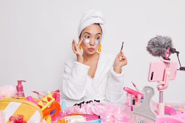 Une jolie femme asiatique tient une brosse professionnelle met des patchs sous les yeux utilise des outils pour les enregistrements de maquillage blog vidéo pour les sites de réseaux sociaux donne des conseils de beauté et de soins de la peau