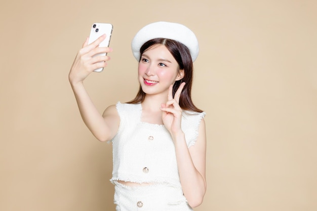 Jolie femme asiatique utilisant un téléphone portable pour un appel vidéo ou un selfie Modèle jolie fille portant un béret blanc et un maquillage naturel sur fond beige Cosmétologie beauté et bien-être spa Chirurgie plastique