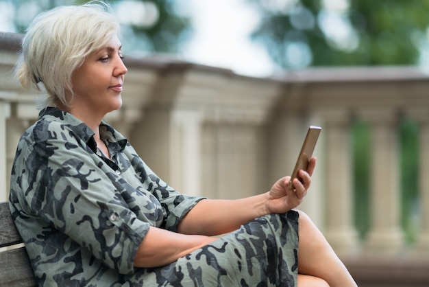 Jolie femme assise sur un banc en plein air la lecture sur son téléphone portable avec un sourire tranquille de plaisir dans une vue latérale rapprochée