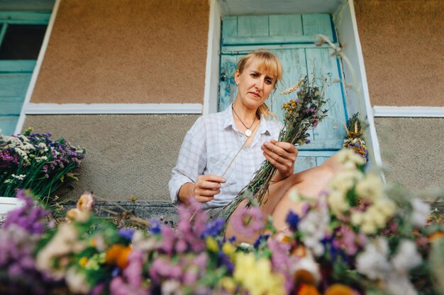 Jolie femme assise dans des fleurs séchées et crée des bouquets de vacances avec un visage sérieux