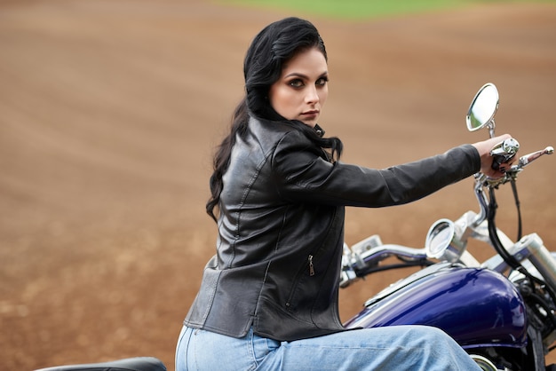 Jolie femme assise sur une moto au coucher du soleil dans un champ.