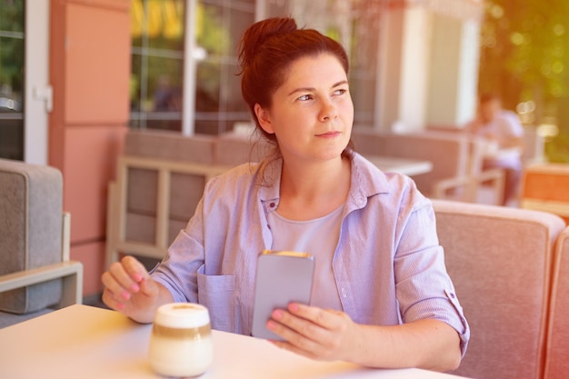 Photo jolie femme assise en plein air avec son smartphone au café avec une tasse de café lumière du soleil sur fond