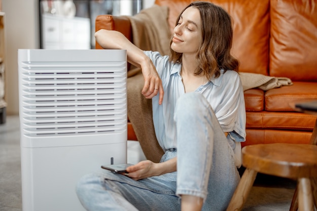 Jolie femme assise près d'un purificateur d'air et d'un appareil hydratant près d'un canapé surveillant la qualité de l'air dans le téléphone. Microclimat de santé à la maison concept.