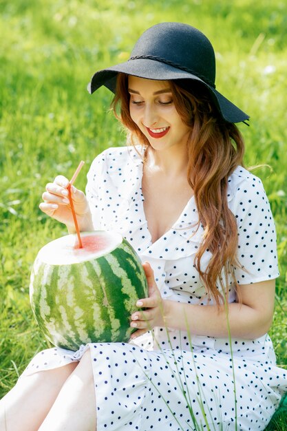 Jolie femme au chapeau noir détient une pastèque entière avec de la paille cocktail.